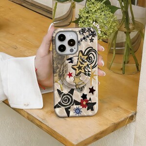 May include: A beige phone case with a collage of star designs in black, gold, red, and blue. The case features a variety of star shapes, including five-pointed stars and starbursts, with a swirling pattern. The case has a camera cutout and a pop-up grip.