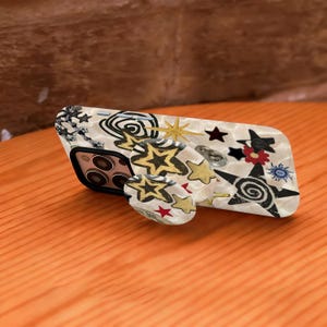 May include: A phone case and matching pop socket with a cream background and a pattern of stars, swirls, and other shapes in black, gold, and red. The phone case has a black border and a camera cutout.