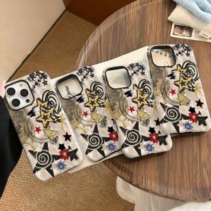 May include: Four phone cases with a decorative design. The cases have a white and gray marble-like background with gold stars, red stars, black and white starbursts, and red and blue flowers. The cases are on a wooden table.