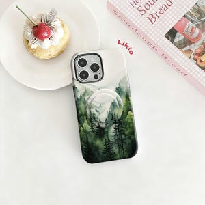May include: A smartphone case with a forest scene design. The case features a watercolor painting of green trees and mountains. A white pop-up grip is attached to the back of the case. The case is on a white surface.