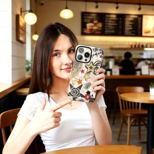 May include: A phone case with a cream-colored background and a collage of stars, moons, and floral designs. The case has a black border and a camera cutout. The design includes gold, red, orange, and black elements.