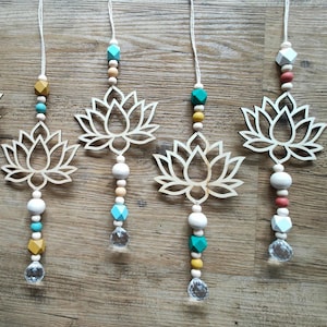Könnte beinhalten: Fünf Holzornamente in Form von Lotusblumen mit Aufhängeschnüren. Jedes Ornament hat ein lasergeschnittenes Lotusdesign, bunte Perlen und einen durchsichtigen Kristallanhänger. Die Perlen sind in verschiedenen Farben gehalten, darunter Weiß, Rosa, Blau und Gelb.