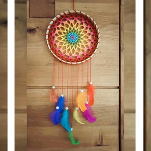 Könnte beinhalten: Ein handgefertigter Traumfänger mit einem kreisförmigen, gehäkelten Netz in Regenbogenfarben. Der Holzreifen ist mit orangefarbenem Faden und bunten Federn in Orange, Blau, Gelb und Lila verziert. Ein dekoratives Element.
