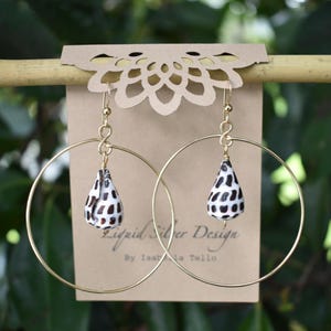 Peut inclure: Boucles d'oreilles créoles dorées avec des pendentifs en forme de goutte d'eau. Les pendentifs sont blancs avec des taches noires. Les boucles sont suspendues à une tige de bambou. Une carte marron avec le texte "Liquid Silver Design" est visible.
