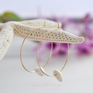 May include: Gold hoop earrings with white, organic-shaped charms. The earrings are displayed on a white starfish against a soft, blurred background of purple flowers. The earrings are delicate and elegant.