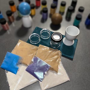 Könnte beinhalten: Eine Auswahl an Bastelbedarf, darunter kleine Gläser, klare ringförmige Objekte und Päckchen mit farbigem Pulver. Ein weißer Eierbecher und ein kleines Glas mit schwarzem Deckel sind ebenfalls zu sehen. Verschiedene kleine Flaschen befinden sich im Hintergrund.
