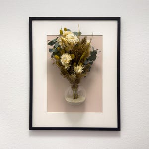 Könnte beinhalten: Ein gerahmtes Blumenarrangement mit einem Strauß getrockneter Blumen in einer kleinen Glasvase. Der Strauß enthält weiße, gelbe und braune Blumen mit grünen Eukalyptusblättern. Der Rahmen ist schwarz mit einem weißen Passepartout und einem rosa Hintergrund.