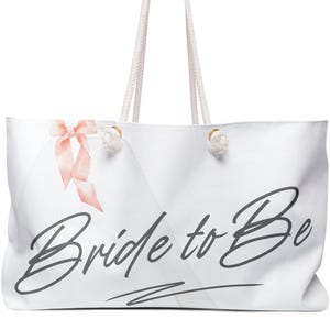 May include: White tote bag with rope handles. The bag features the text "Bride to Be" in a script font, a pink bow, and gold-colored accents. The bag is designed for carrying items.