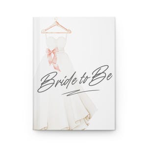 May include: A white journal with a watercolor illustration of a wedding dress on a hanger, accented with a pink bow. The words "Bride to Be" are written in cursive across the front.