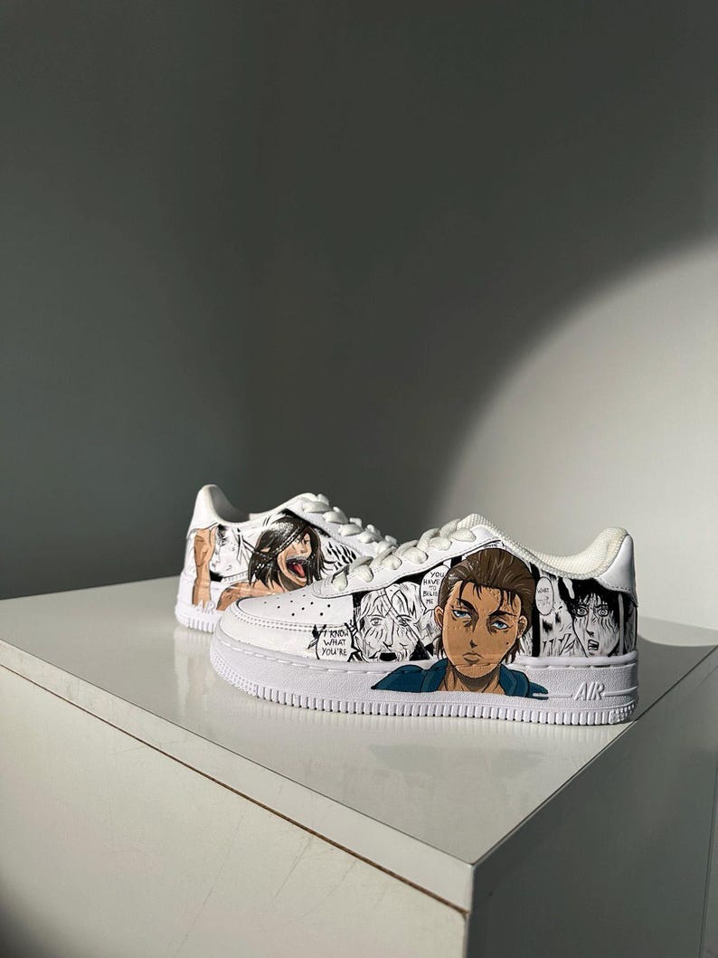 K&ouml;nnte beinhalten: Wei&szlig;e Sneaker mit einem individuellen Design, das Figuren aus dem Anime Attack on Titan zeigt. Das Design umfasst Schwarzwei&szlig;illustrationen der Figuren, darunter Eren Yeager, Mikasa Ackerman und Armin Arlert. Die Sneaker haben das Wort "AIR" an der Seite.