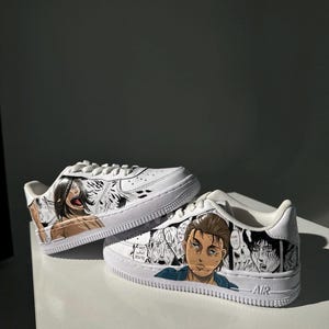 K&ouml;nnte beinhalten: Wei&szlig;e Sneaker mit einem individuellen Design, das Charaktere aus dem Anime Attack on Titan zeigt. Das Design beinhaltet Schwarzwei&szlig;illustrationen der Charaktere, wobei ein Charakter farbig ist. Der Text "AIR" ist an der Seite des Schuhs sichtbar.