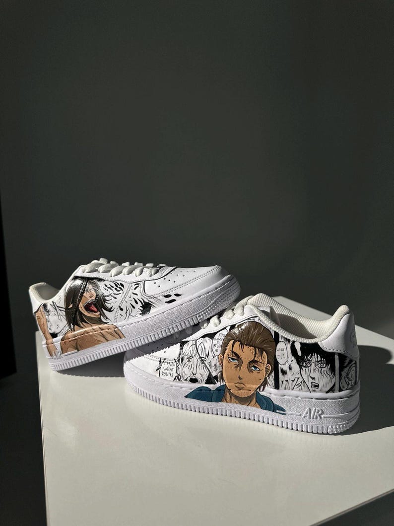 K&ouml;nnte beinhalten: Wei&szlig;e Sneaker mit einem individuellen Design, das Charaktere aus dem Anime Attack on Titan zeigt. Das Design beinhaltet Schwarzwei&szlig;illustrationen der Charaktere, wobei ein Charakter farbig ist. Der Text "AIR" ist an der Seite des Schuhs sichtbar.