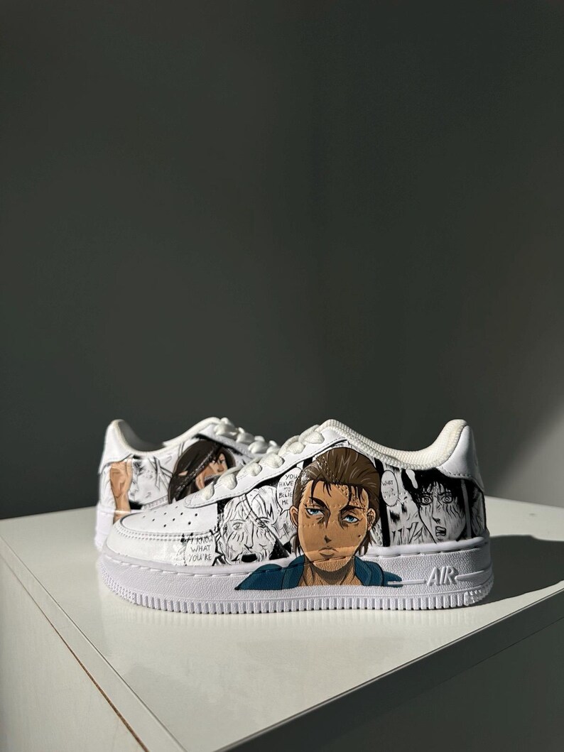 K&ouml;nnte beinhalten: Wei&szlig;e Sneaker mit einem individuellen Design, das Charaktere aus dem Anime Attack on Titan zeigt. Das Design beinhaltet Schwarzwei&szlig;-Manga-Panels und eine Nahaufnahme eines Gesichts. Das Wort "AIR" ist an der Seite des Schuhs aufgedruckt.