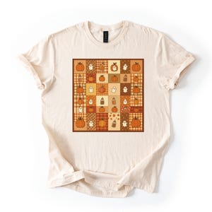 Puede incluir: Una camiseta de color crema con un gráfico cuadrado central compuesto por una cuadrícula de cuadrados más pequeños. La cuadrícula muestra un patrón repetitivo de calabazas naranjas, fantasmas blancos y texturas de cuadros marrones.