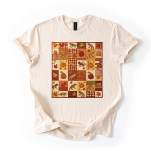 Puede incluir: Una camiseta de manga corta de color crema con un gráfico cuadrado central compuesto por una cuadrícula de patchwork. La cuadrícula muestra ilustraciones alternas de lagartos, calabazas y hojas de otoño en tonos naranja, marrón y rojo.