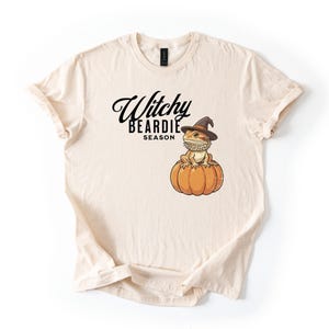 Puede incluir: Una camiseta de color crema con un gráfico de un dragón barbudo que lleva un sombrero de bruja y está sentado sobre una calabaza. El texto "Witchy BEARDIE SEASON" está impreso en negro sobre la ilustración. La camiseta tiene mangas cortas.