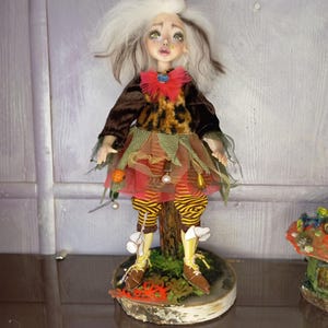 May include: A handcrafted doll with a whimsical design. The doll features a white and brown hairstyle, a brown velvet top, and a dress with orange, red, and green accents. The doll stands on a wooden base with brown shoes and yellow and brown striped leggings.