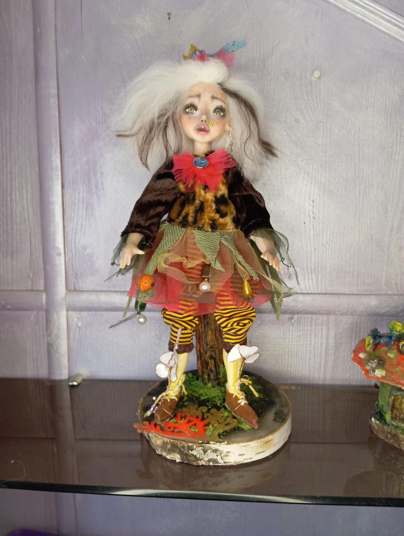 May include: A handcrafted doll with a whimsical design. The doll features a white and brown hairstyle, a brown velvet top, and a dress with orange, red, and green accents. The doll stands on a wooden base with brown shoes and yellow and brown striped leggings.