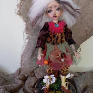 May include: A handcrafted art doll with white and brown hair, wearing a burgundy velvet jacket, leopard print bodice, and layered skirt. The doll has a whimsical crown and is posed on a wooden base with painted details. The doll's outfit includes striped leggings and brown shoes.