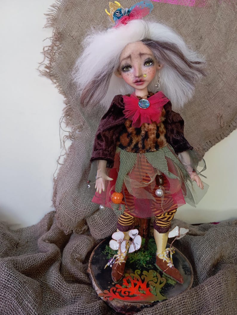May include: A handcrafted art doll with white and brown hair, wearing a burgundy velvet jacket, leopard print bodice, and layered skirt. The doll has a whimsical crown and is posed on a wooden base with painted details. The doll's outfit includes striped leggings and brown shoes.