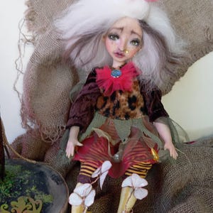 May include: A handcrafted doll with a whimsical design. The doll features a white and brown wig, a small gold crown, and a red ruffled collar with a blue gem. It wears a leopard print top, a layered skirt, and striped leggings with flower accents.