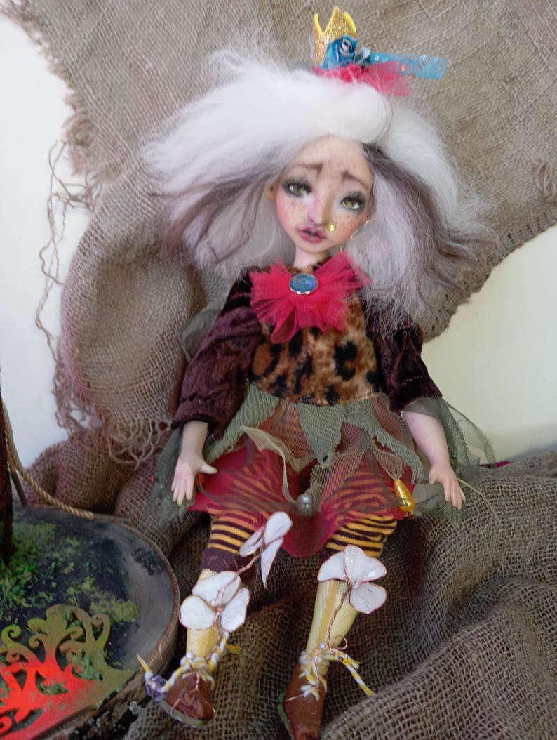 May include: A handcrafted doll with a whimsical design. The doll features a white and brown wig, a small gold crown, and a red ruffled collar with a blue gem. It wears a leopard print top, a layered skirt, and striped leggings with flower accents.
