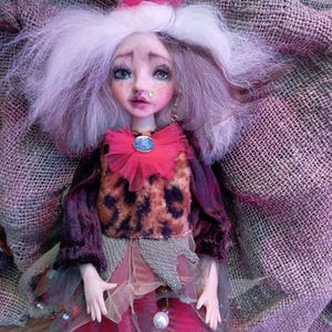May include: A handmade art doll with a whimsical design. The doll features a detailed face with painted features, white hair, and a small crown. It wears a layered outfit with a leopard print top, a pink tulle skirt, and green leggings. The doll is posed on a textured fabric.
