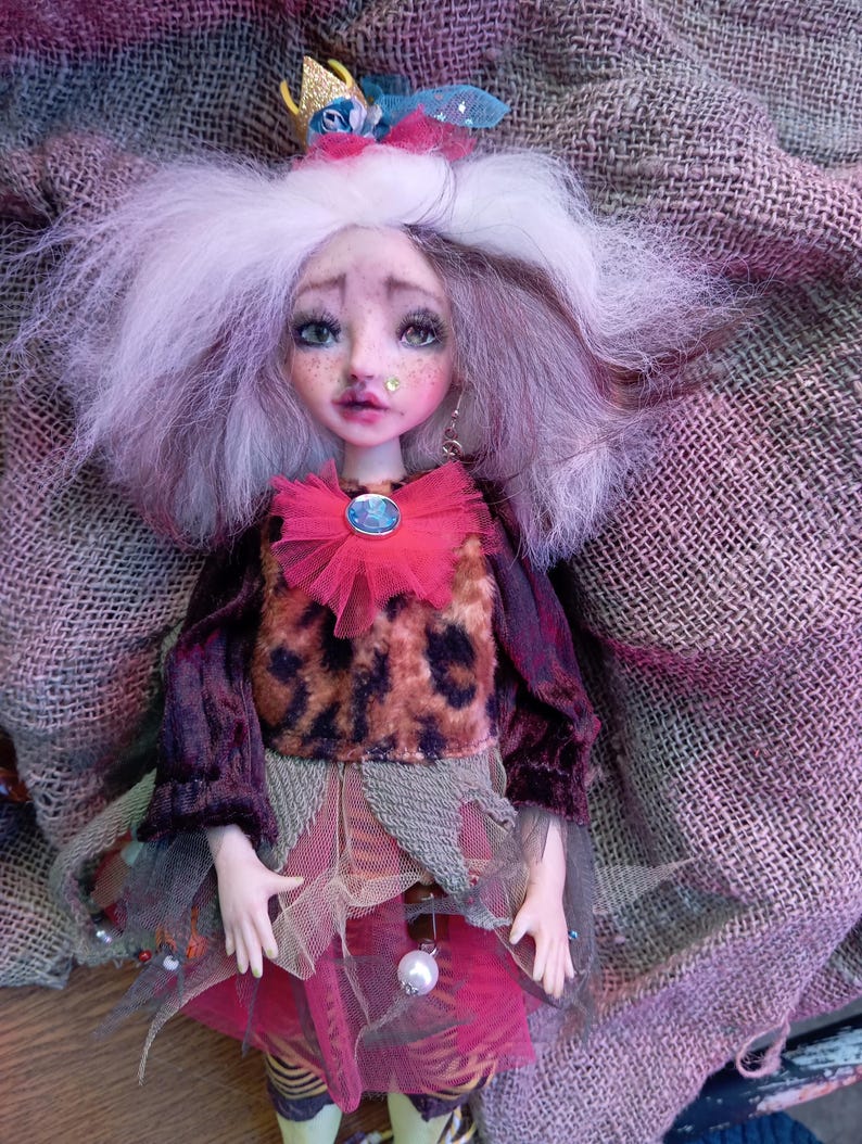 May include: A handmade art doll with a whimsical design. The doll features a detailed face with painted features, white hair, and a small crown. It wears a layered outfit with a leopard print top, a pink tulle skirt, and green leggings. The doll is posed on a textured fabric.