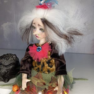 May include: A handmade art doll with a whimsical design. The doll has white and brown hair, a long nose, and is wearing a crown, a red and blue jeweled collar, and a multi-layered dress. The doll's outfit includes a brown jacket, a leopard print top, and striped leggings.