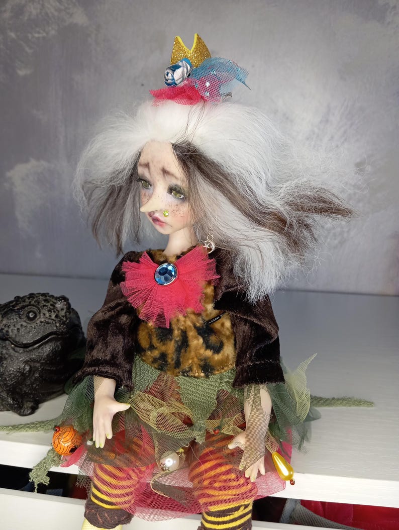May include: A handmade art doll with a whimsical design. The doll has white and brown hair, a long nose, and is wearing a crown, a red and blue jeweled collar, and a multi-layered dress. The doll's outfit includes a brown jacket, a leopard print top, and striped leggings.