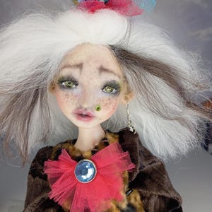 May include: A handmade doll with a whimsical design. The doll has white and brown hair, green eyes, and a red tulle bow with a blue gem. It wears a brown jacket and a gold crown. The doll has a unique, artistic appearance.