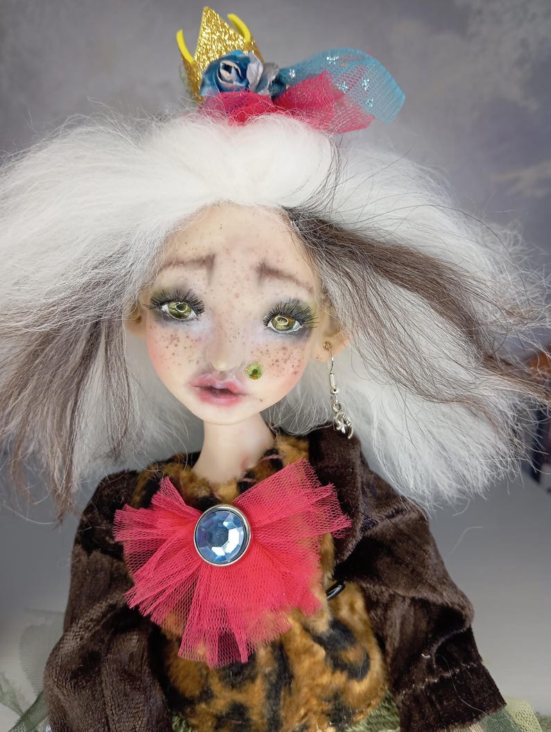 May include: A handmade doll with a whimsical design. The doll has white and brown hair, green eyes, and a red tulle bow with a blue gem. It wears a brown jacket and a gold crown. The doll has a unique, artistic appearance.