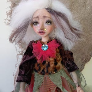 May include: A handcrafted art doll with a whimsical design. The doll features white and brown hair, green eyes, and a detailed face with freckles. It wears a velvet jacket, a leopard print top, and a layered skirt with a red flower and a blue gem.