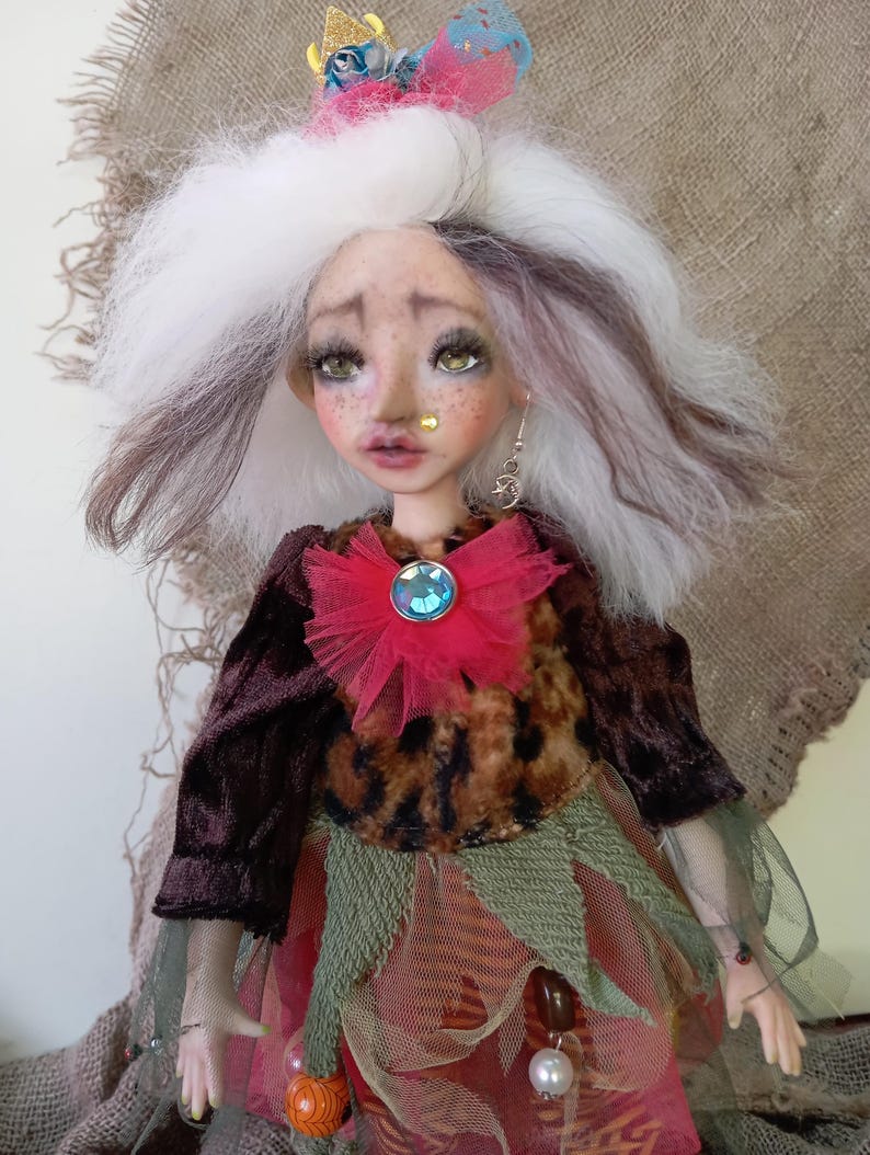 May include: A handcrafted art doll with a whimsical design. The doll features white and brown hair, green eyes, and a detailed face with freckles. It wears a velvet jacket, a leopard print top, and a layered skirt with a red flower and a blue gem.