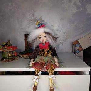 May include: A handmade doll with a whimsical design. The doll has white hair, a red bow, and a crown. It wears a brown jacket, a leopard print top, and striped pants. The doll is sitting on a white surface, with a mushroom house and other decorative items in the background.