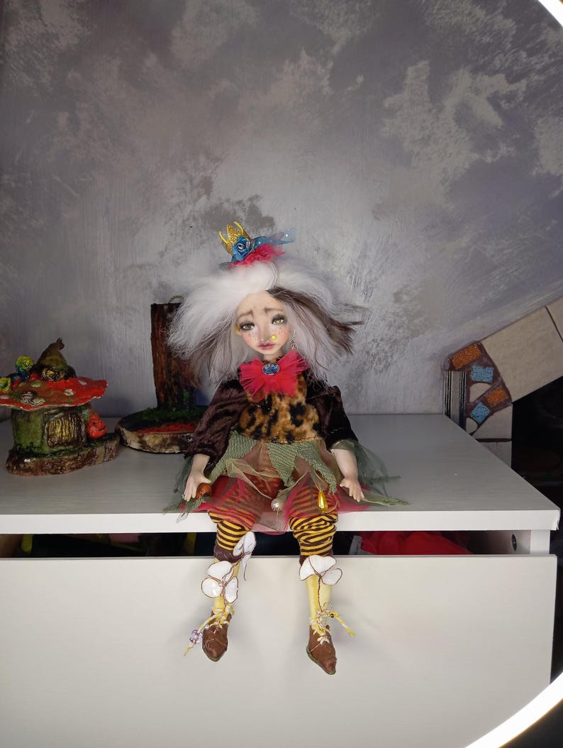 May include: A handmade doll with a whimsical design. The doll has white hair, a red bow, and a crown. It wears a brown jacket, a leopard print top, and striped pants. The doll is sitting on a white surface, with a mushroom house and other decorative items in the background.