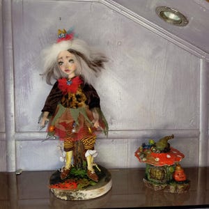 May include: A handcrafted doll with white and brown hair, wearing a brown jacket, leopard print top, and colorful skirt. The doll stands on a wooden base. Beside it is a miniature mushroom house with a jack-o'-lantern.