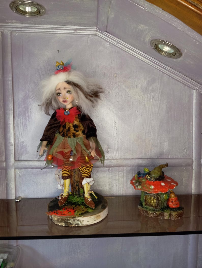 May include: A handcrafted doll with white and brown hair, wearing a brown jacket, leopard print top, and colorful skirt. The doll stands on a wooden base. Beside it is a miniature mushroom house with a jack-o'-lantern.