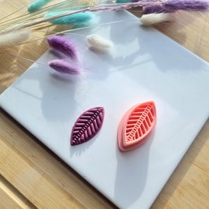 May include: Two leaf-shaped silicone molds, one pink and one purple, on a white surface. The molds are designed to create detailed leaf shapes.
