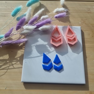 May include: Two pairs of earrings, one pair is pink and the other is blue. Both pairs are shaped like a chevron with a pointed tip.