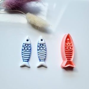 May include: Three fish-shaped objects. Two are white with blue detailing, and one is pink. The fish have a simple, stylized design with fins and scales. The objects are on a white surface, with dried flowers in the background.