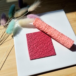 May include: A pink and red rolling pin and a square textured stamp with a floral design. Both tools are used for decorating fondant or clay.