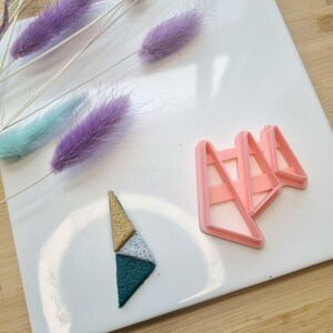 May include: A pink plastic triangle cookie cutter with three different sized triangles. A green, white, and gold triangle shaped cookie is on a white surface.