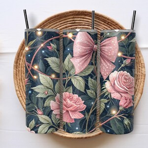 May include: Three stainless steel tumblers with black straws, featuring a floral design with pink roses, green leaves, and a pink bow on a dark blue background. The tumblers are set in a woven basket.