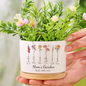 Puede incluir: Maceta de cerámica blanca con base de madera. La maceta tiene un diseño floral y los nombres Candace, Beulah, Karen, Agatha, Queena y Nirma escritos en ella. El texto "Mom's Garden - best mom ever" también está impreso en la maceta.