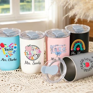 May include: Five cylindrical tumblers with clear lids and grey sliding closures. The tumblers are light blue, white, pink, black, and grey, each featuring colorful teacher-themed graphics, apples, and personalized names like Mrs. Clarke and Mrs. Sandy.