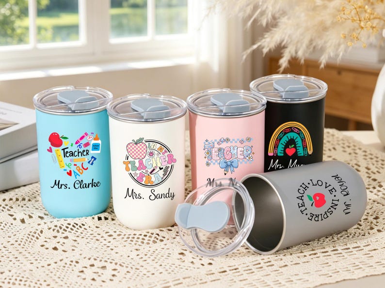 May include: Five cylindrical tumblers with clear lids and grey sliding closures. The tumblers are light blue, white, pink, black, and grey, each featuring colorful teacher-themed graphics, apples, and personalized names like Mrs. Clarke and Mrs. Sandy.