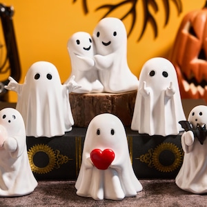 Op de afbeelding: Een verzameling witte keramische spookfiguren met zwarte ogen, sommige met een hart, een pompoen of vleermuisvleugels. Een gesneden oranje pompoen en andere Halloween-decoraties staan op de achtergrond.