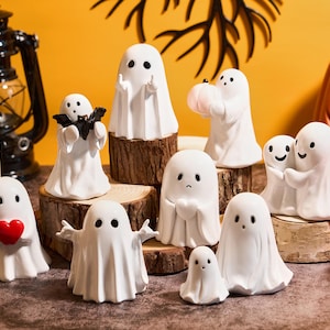 Könnte beinhalten: Eine Sammlung weißer Keramik-Geisterfiguren mit verschiedenen Posen und Accessoires, darunter ein Herz, eine Fledermaus und ein Kürbis. Die Halloween-Dekorationen werden auf Holzplattformen und einem Tisch präsentiert.