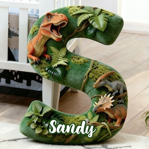 May include: A green, dinosaur-themed letter "S" with the name "Sandy" in white script. The letter is decorated with various dinosaur figurines, ferns, and moss. An orange T-Rex is featured at the top.